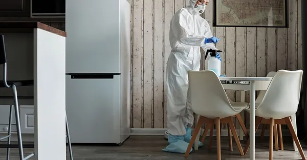 Person in hazmat suit disinfecting a modern kitchen table with spray bottle for hygiene. cleaner with spray bottle during disinfection of apartment