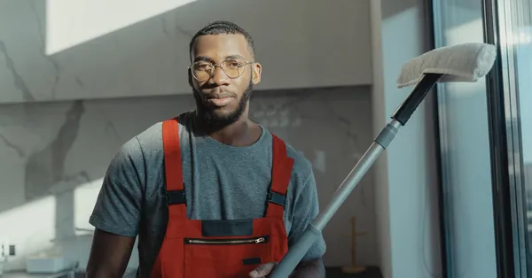 A professional cleaner holding a tool indoors, dressed in red overalls with natural light. a bearded man holding a cleaning tool