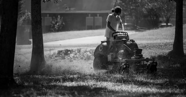 A person operating a lawnmower in a shaded, grassy yard, in black and white. black and white image of person mowing lawn outdoors